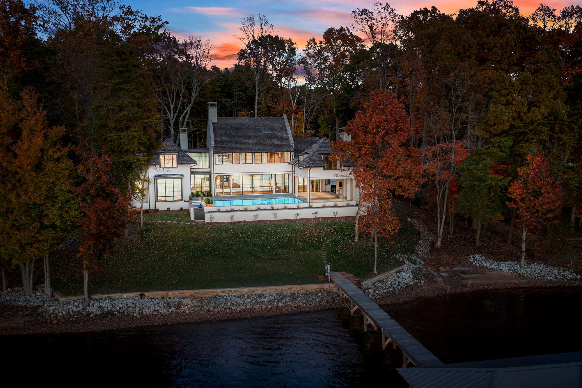 A large modern house with a swimming pool on a hillside overlooking water, surrounded by autumn trees, during sunset.