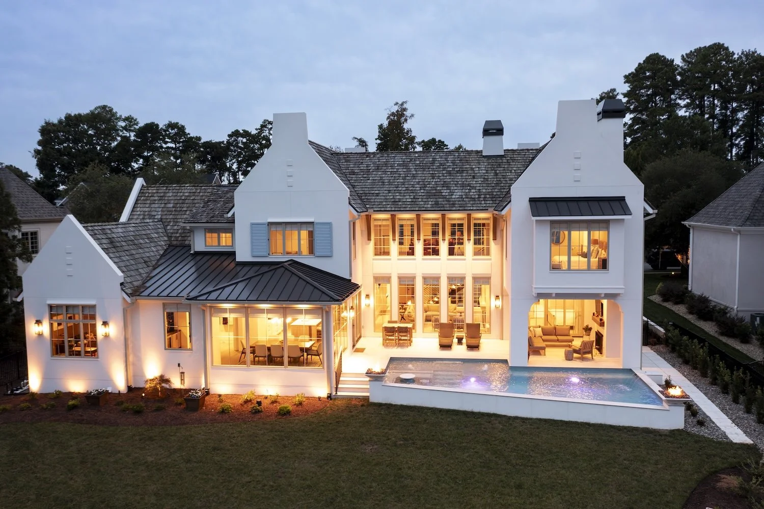 Large white house at dusk with lights on, a backyard pool, outdoor seating, and a landscaped yard.
