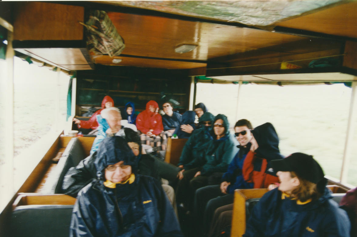 A group of people wearing raincoats and sunglasses riding on the back of a truck, with some looking out the windows and others talking or smiling.