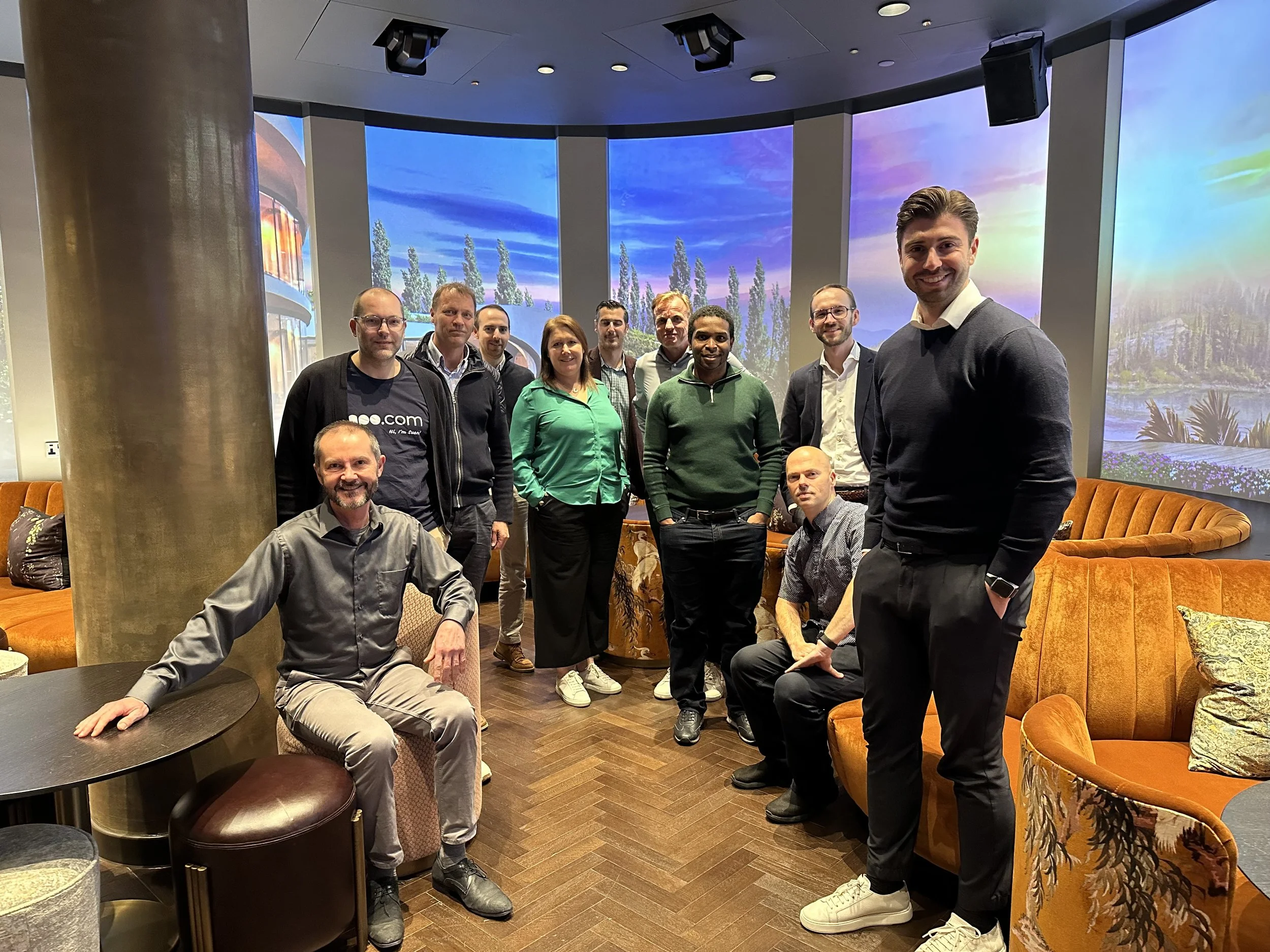 Group of eleven people posing indoors with curved wall screens displaying scenic landscapes in the background, sitting and standing on wooden flooring with orange and patterned sofas.