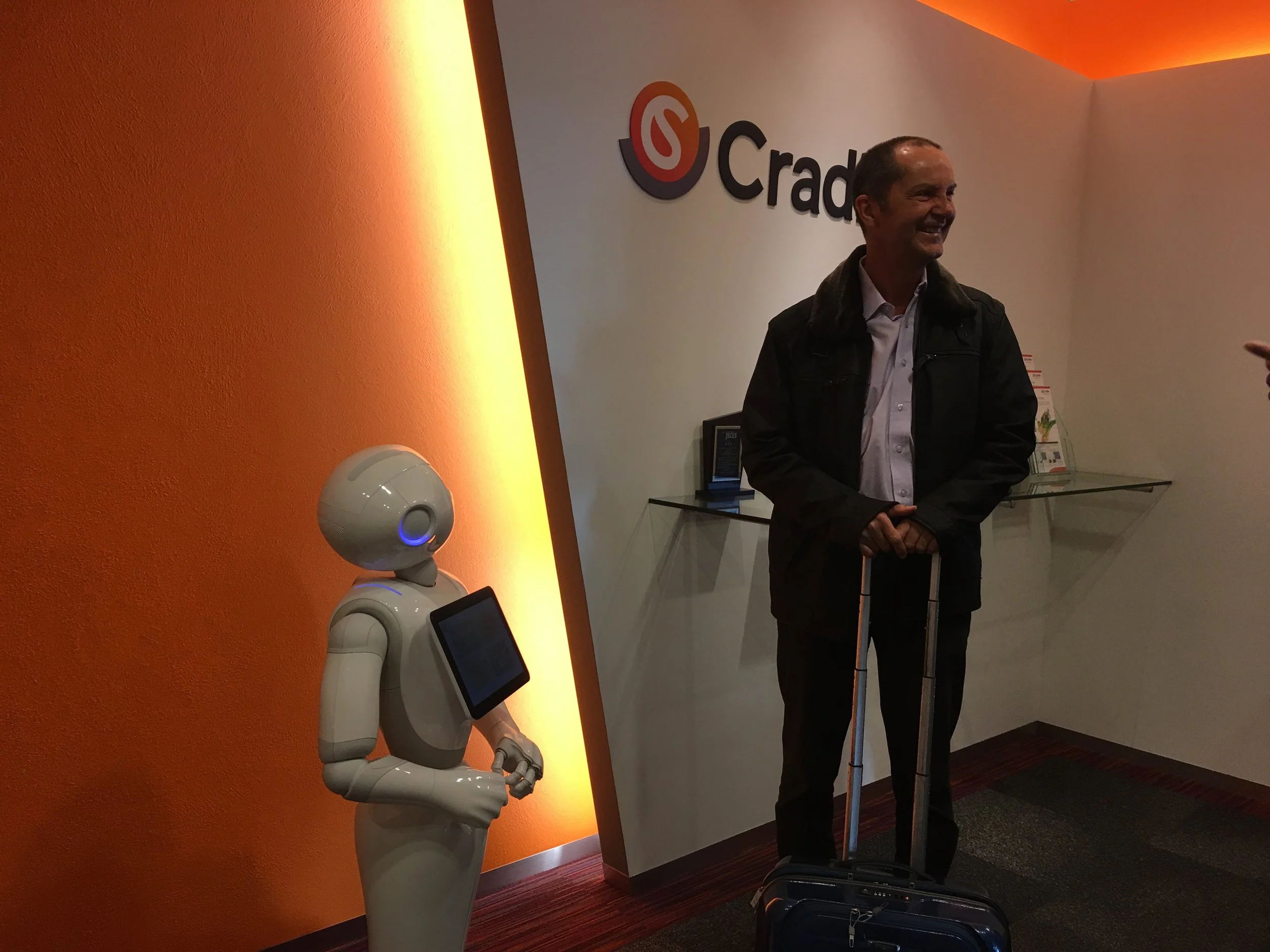 A man standing with a rolling suitcase in an office lobby near a humanoid robot with a tablet on its chest. The background includes an orange and white wall with a company logo that says 'Cradle'.