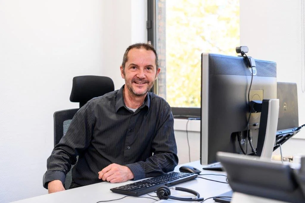 A man with a beard and short hair is sitting at a desk in an office, smiling, with a computer, keyboard, mouse, and headset in front of him. There is a window behind him showing trees outside.