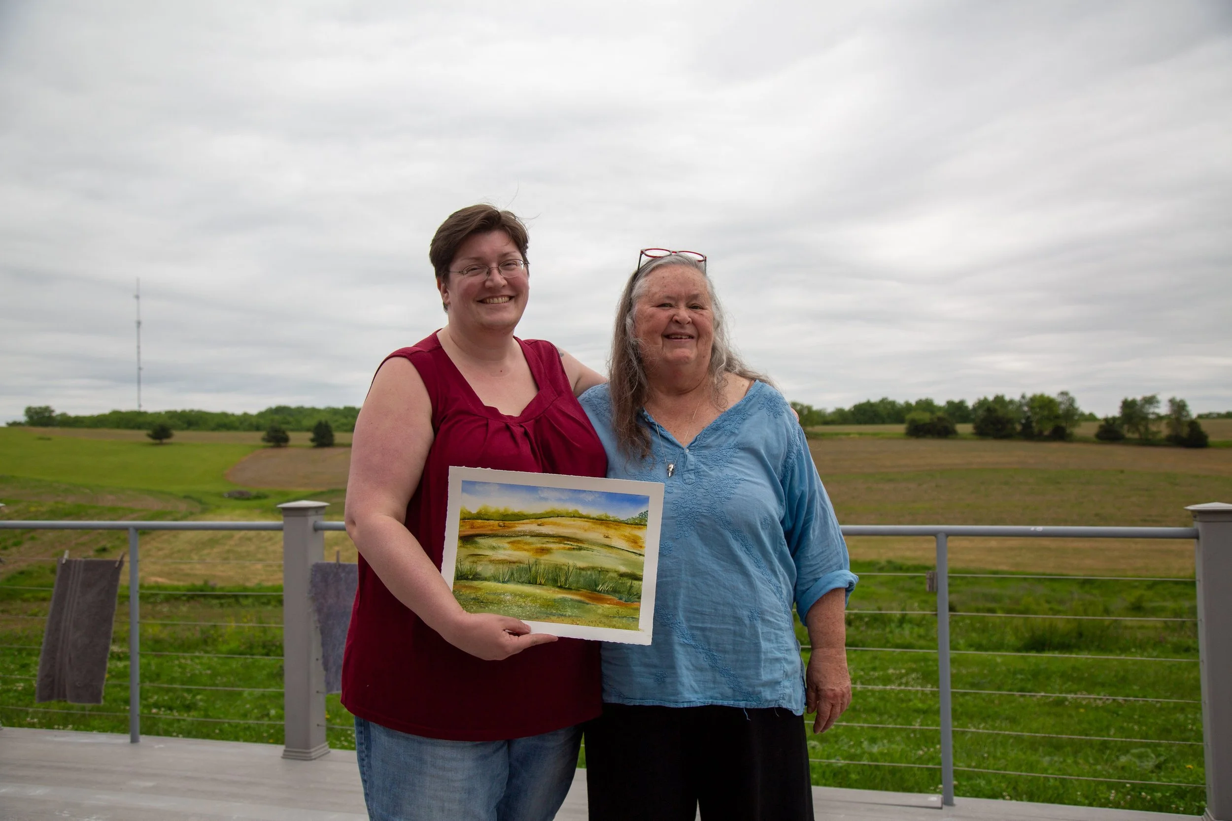 Stacy and Helen standing next to each other in front of a cornfield while Stacy holds a painting of harvested fields. 