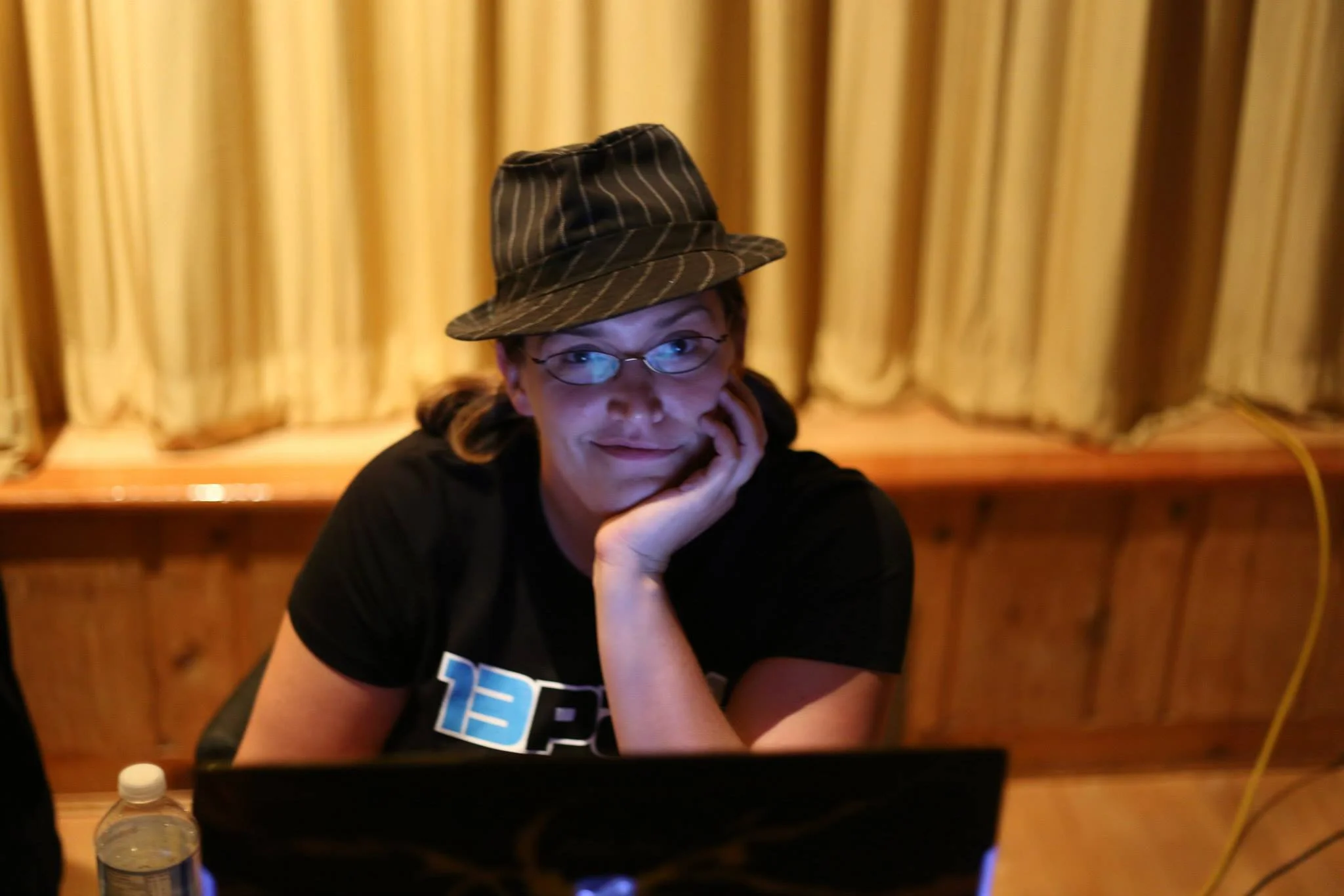 Stacy sitting infront of an open laptop, her head in her hand, the screen reflecting in her glasses, wearing her fedora hat
