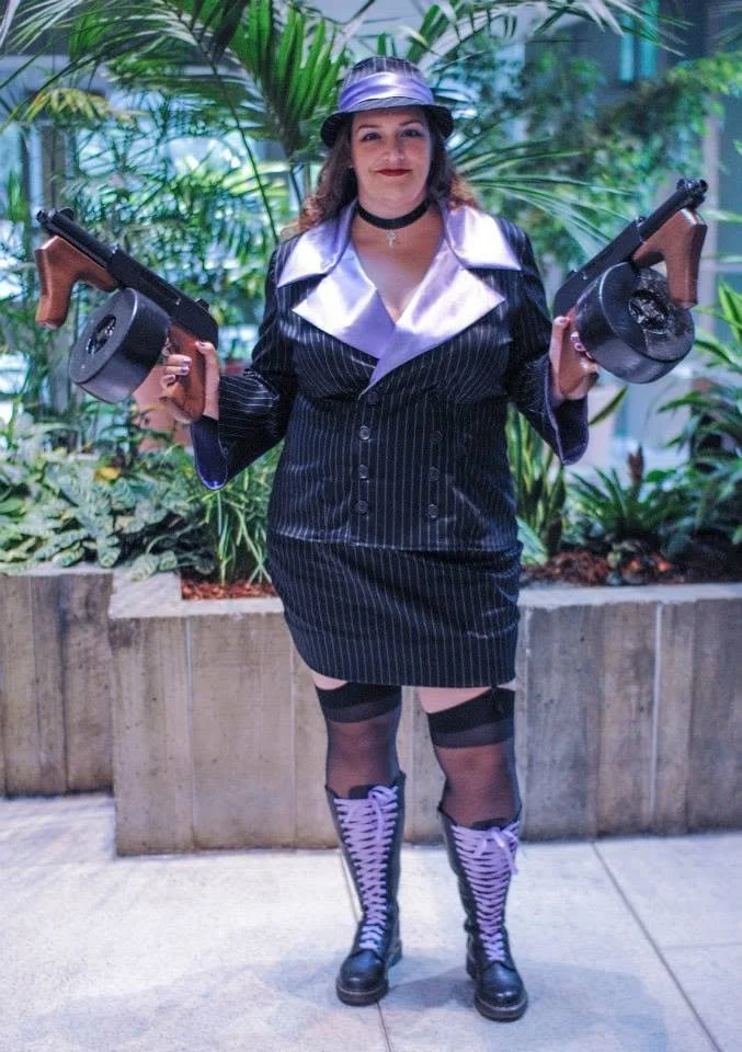 Stacy in Mafia Miss Fortune cosplay, wearing a pinstripe suit with a fedora and holding two fake tommies