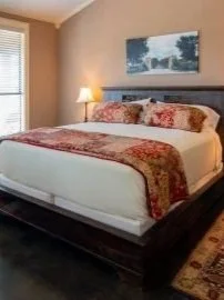 A neatly made bed with floral bedding and pillows in a bedroom with a beige wall, a window with blinds, and a table lamp.