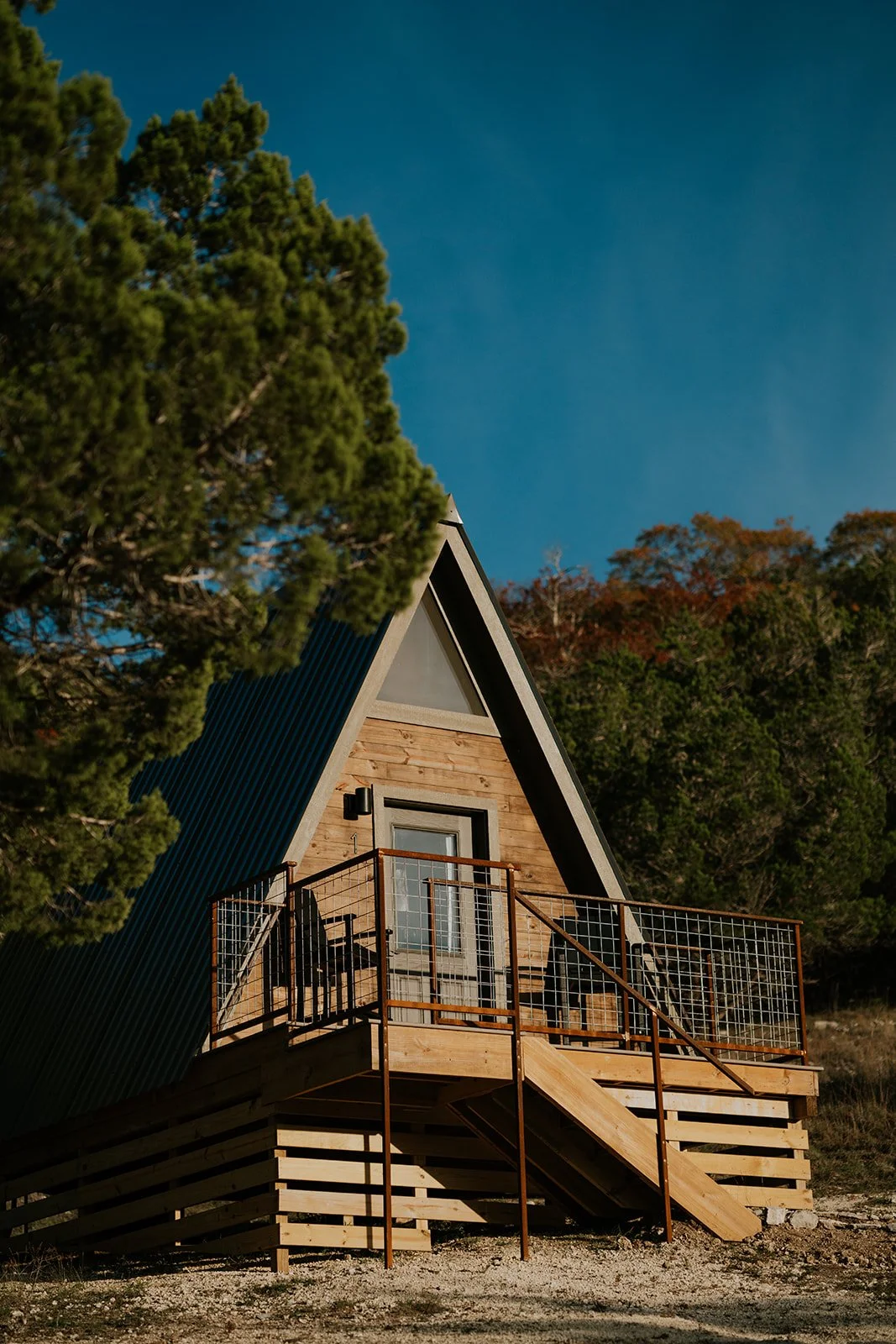 Aerial A-frame cabin with a wooden deck, metal railing, and glass door, surrounded by trees under a clear blue sky.