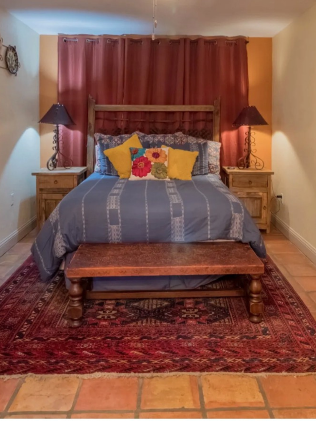 A bedroom with a bed, two wooden nightstands with lamps, red curtains, and a decorative rug.