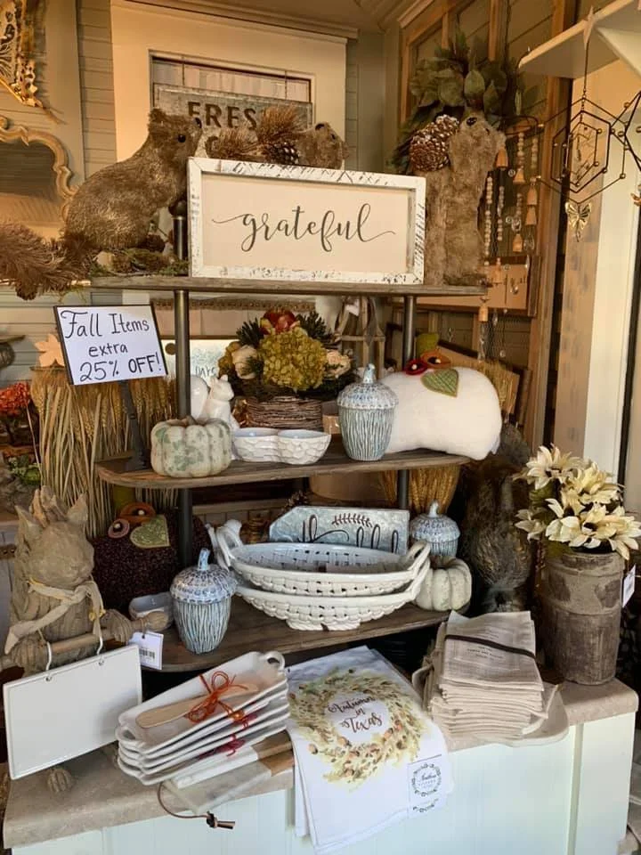 Display of fall home decor items including woven pumpkins, books, baskets, vases, and scented candles on a multi-tiered shelf with a sign reading 'grateful' and a sign offering 25% off on fall items.