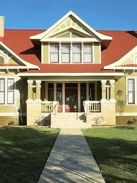 Front view of a yellow, two-story house with a red roof, front porch with steps, and a walkway leading to the door. The house has multiple windows and decorative trim.