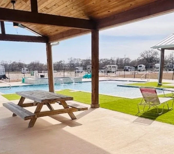 Outdoor swimming pool area with a picnic table, lounge chairs, and green artificial grass under a wooden shelter, surrounded by RVs and a fence.