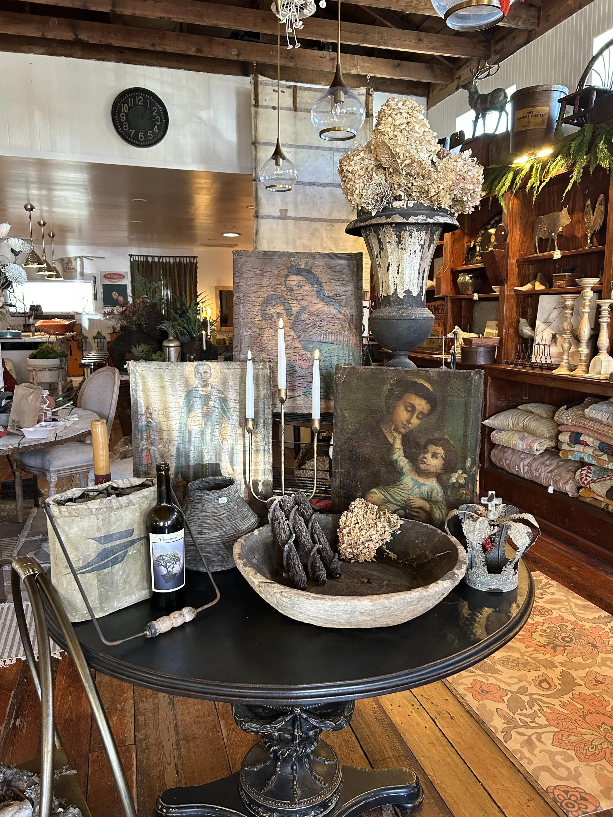A black round table with vintage paintings, dried flowers, candles, and decorative items inside a rustic store.