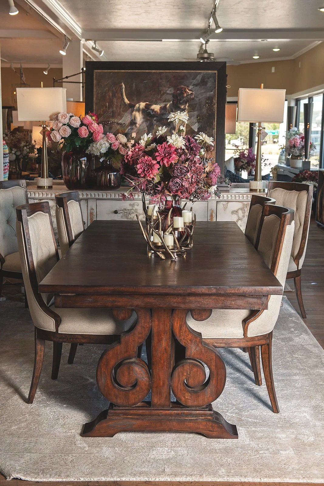A dining area with a dark wooden table surrounded by six upholstered chairs, a large floral centerpiece, and lamps on side tables in the background. There's a painting of a dog behind the table and decorative vases with flowers on a sideboard.