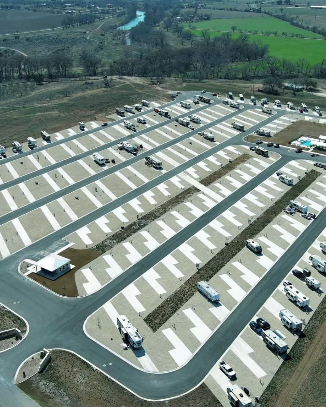 A large parking lot under construction with many parked RVs and trucks, some parking spaces marked, near a building with a pool, surrounded by open land and trees, in a rural area.