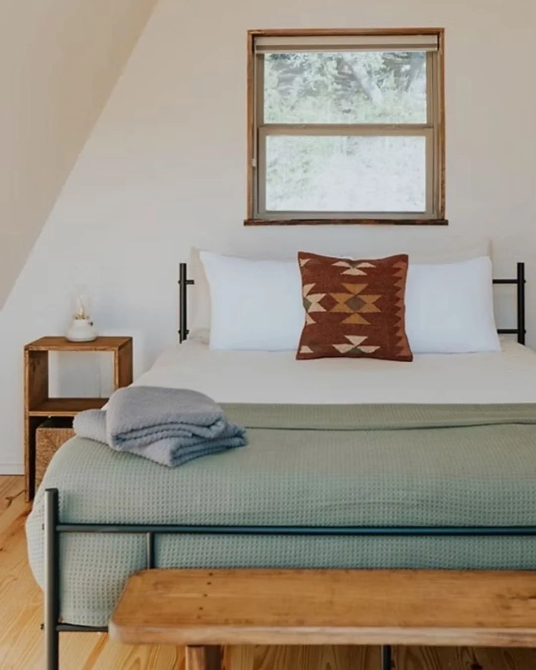 A cozy bedroom with a metal bed frame, light green bedspread, and a decorative pillow with a geometric pattern. Folded gray and white towels are on the bed. There is a wooden nightstand with a small white vase next to the bed. A window with a wooden frame shows greenery outside.