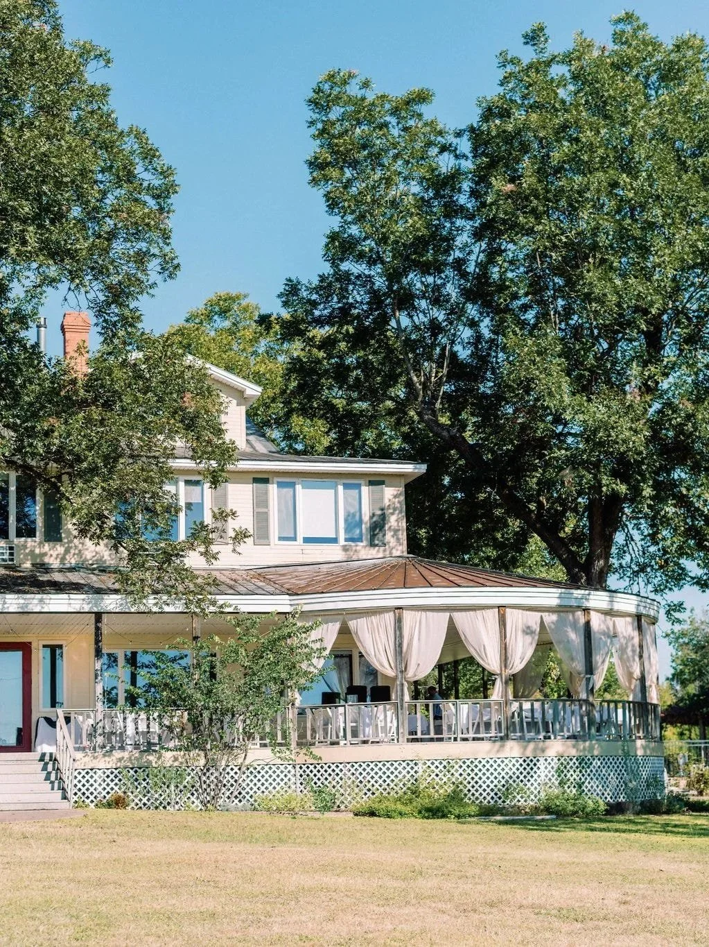 A two-story house with a large wrap-around porch covered by beige curtains, surrounded by lush green trees and a well-maintained lawn, under a clear blue sky.