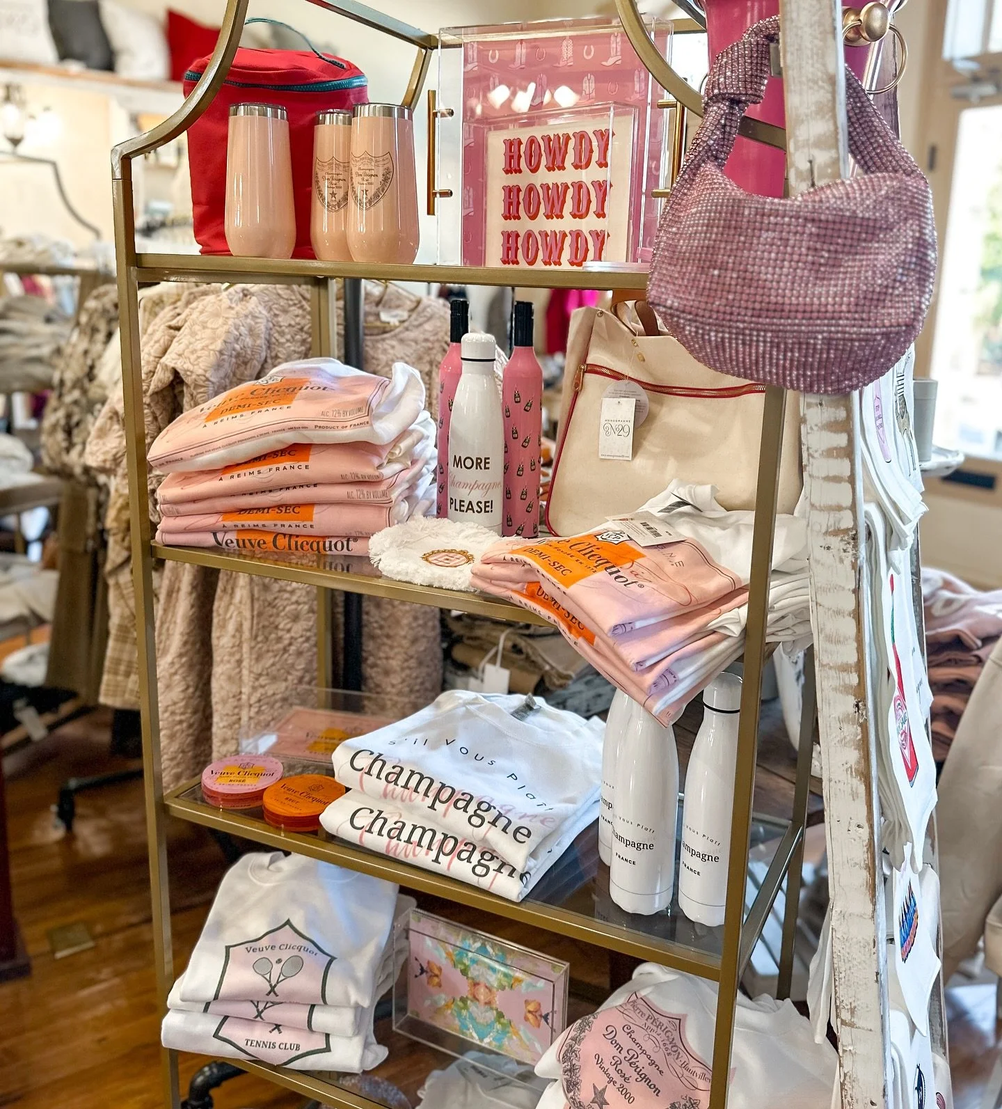 Display shelf with pink and white decorative items, towels, water bottles, and bags, with some items labeled 'Champagne' and 'Veuve Clicquot' in a retail store.