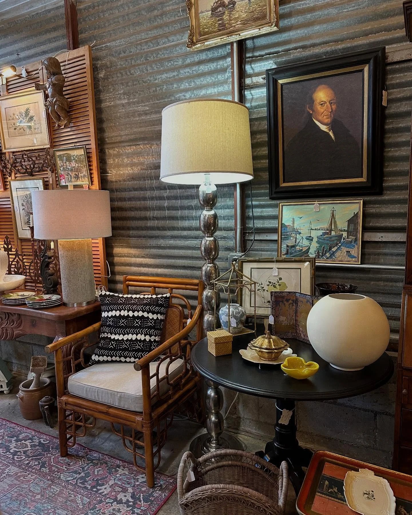 A corner of an antique shop with various vintage furniture, lamps, artwork, and decorative items displayed against a corrugated metal wall.
