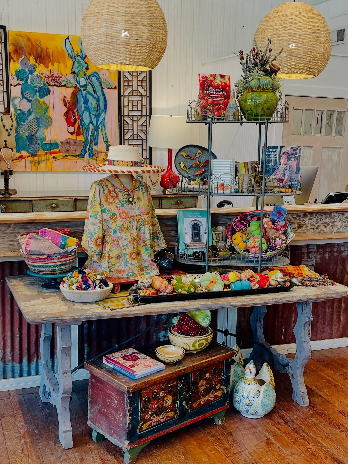 A colorful display of clothing, bowls, and decorative items on a rustic wooden table in an eclectic store. Includes a mannequin dressed in a floral dress with a wide-brimmed hat, baskets of handmade items, a three-tiered metal shelf with decorative balls and figurines, and vibrant artwork on the wall.