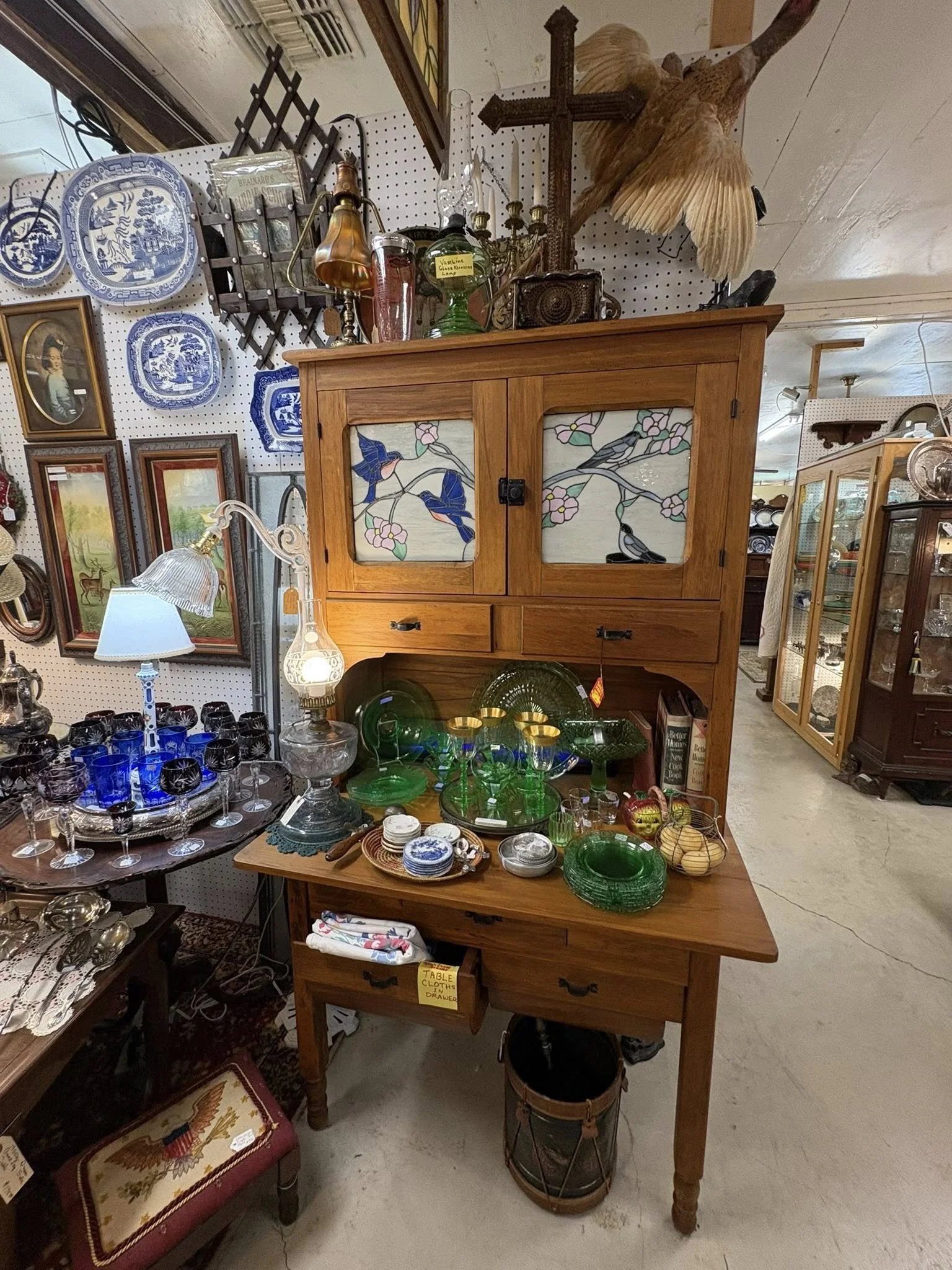 A wooden display cabinet with stained glass panels featuring bird and flower designs, situated in an antique shop among various glassware, ceramics, and decorative items.