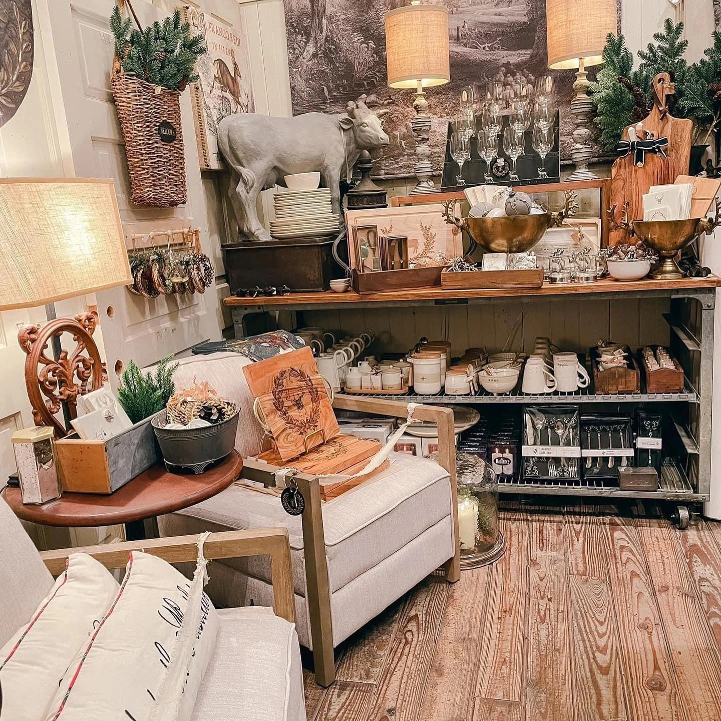 An interior view of a cozy store with home decor and kitchenware, featuring a beige couch with pillows, a wooden side table with a lamp, a shelving unit with dishes, mugs, and silverware, surrounded by decorative items including a cow sculpture, lamps, bowls, plants, and artwork.