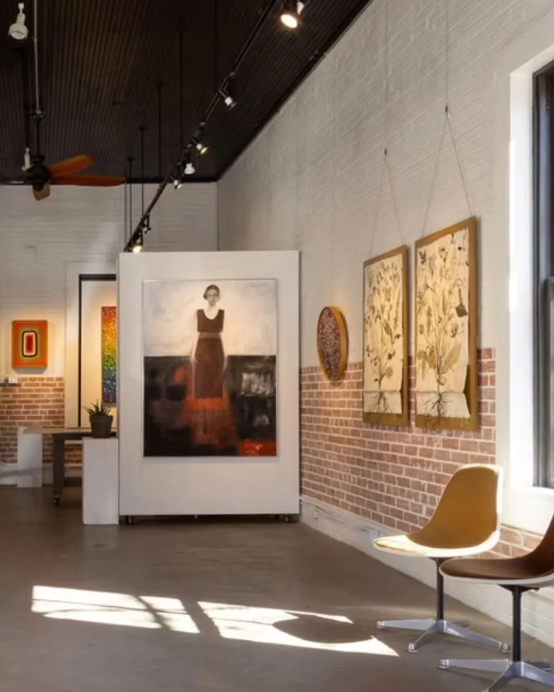 Interior of an art gallery with paintings on white and brick walls, sunlight streaming through a window, and modern chairs on the floor.
