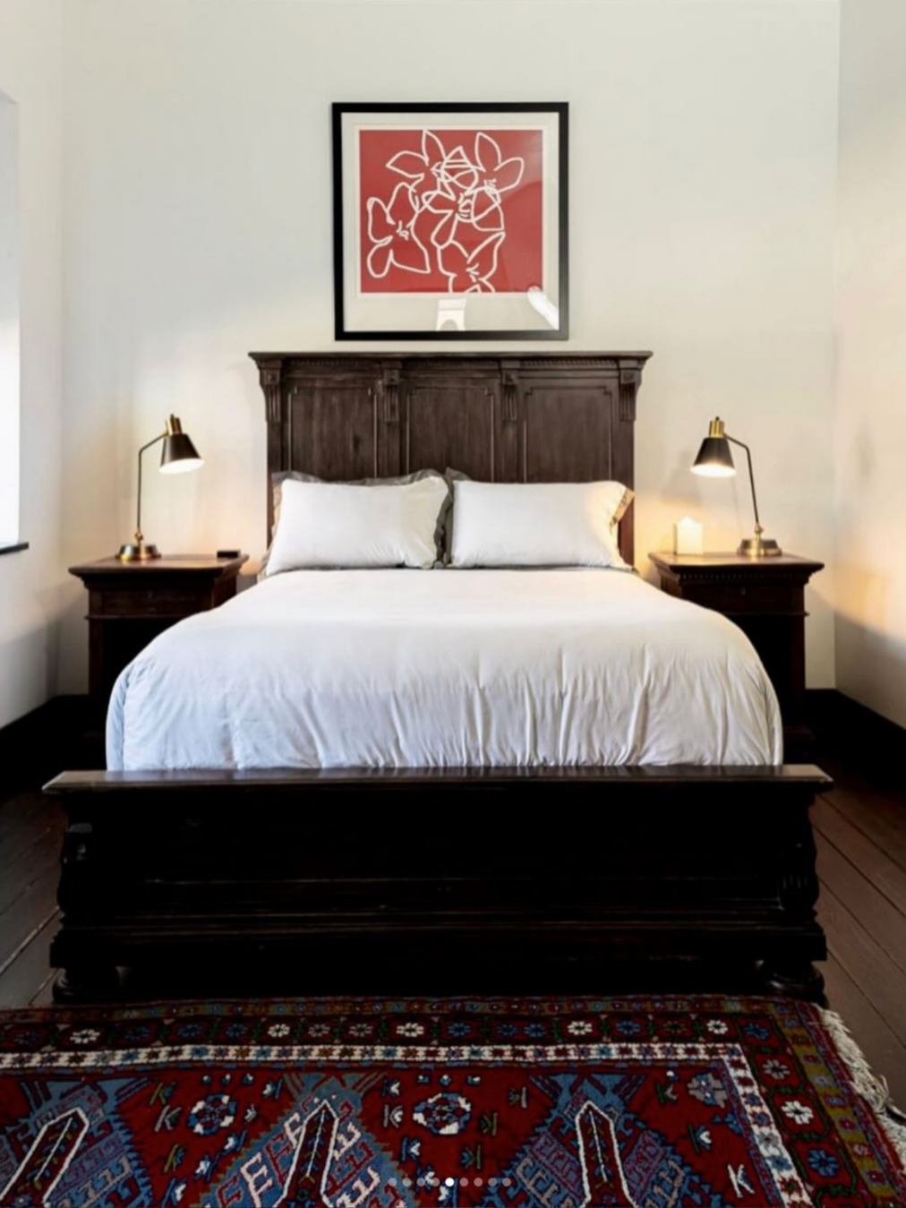 A bedroom with a wooden bed frame, white bedding, two pillows, and two matching bedside tables with lamps. A framed abstract line art picture hangs above the bed, and a colorful patterned rug is partially visible at the foot of the bed.