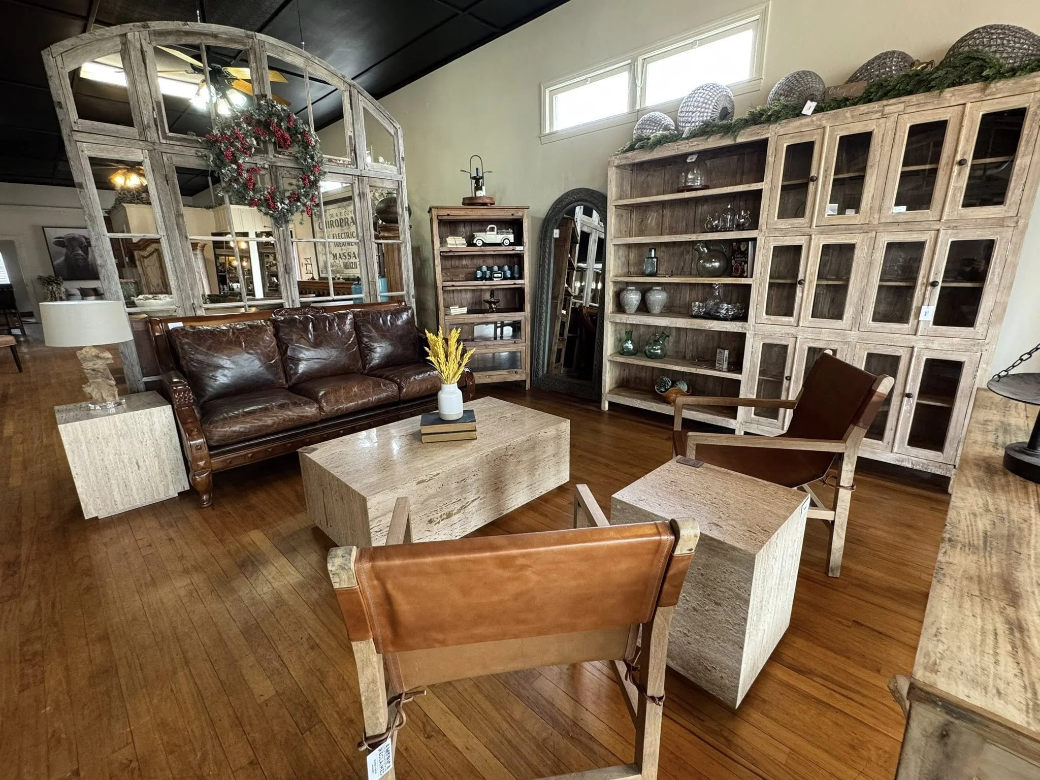 Living room with wooden furniture, a brown leather sofa, armchairs, a coffee table, and large wooden cabinets filled with glassware and decorative items. A wreath decorated with red berries hangs on a decorative window frame.