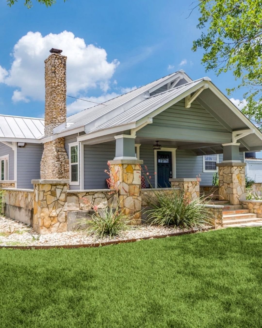 Front view of a house with gray siding, stone accents, a porch, a chimney made of small stones, and a well-maintained lawn under a partly cloudy sky.