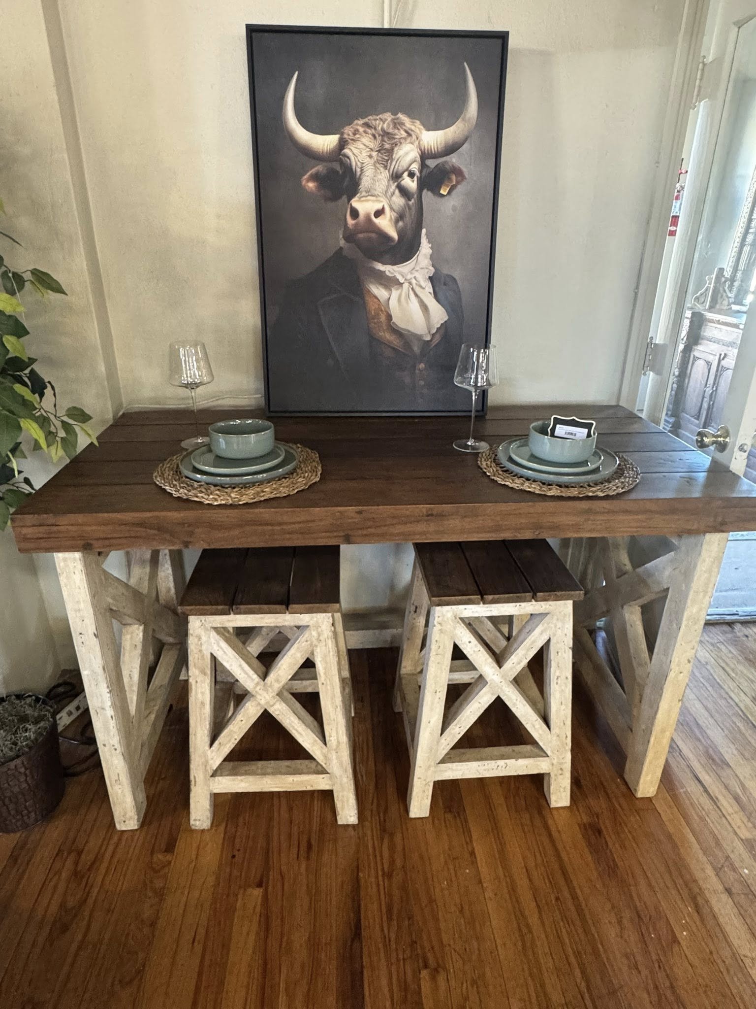A dining table with two place settings, a large framed painting of a cow in a suit with a ruffled shirt, and two wine glasses inside a room with wooden floors and a door to the right.