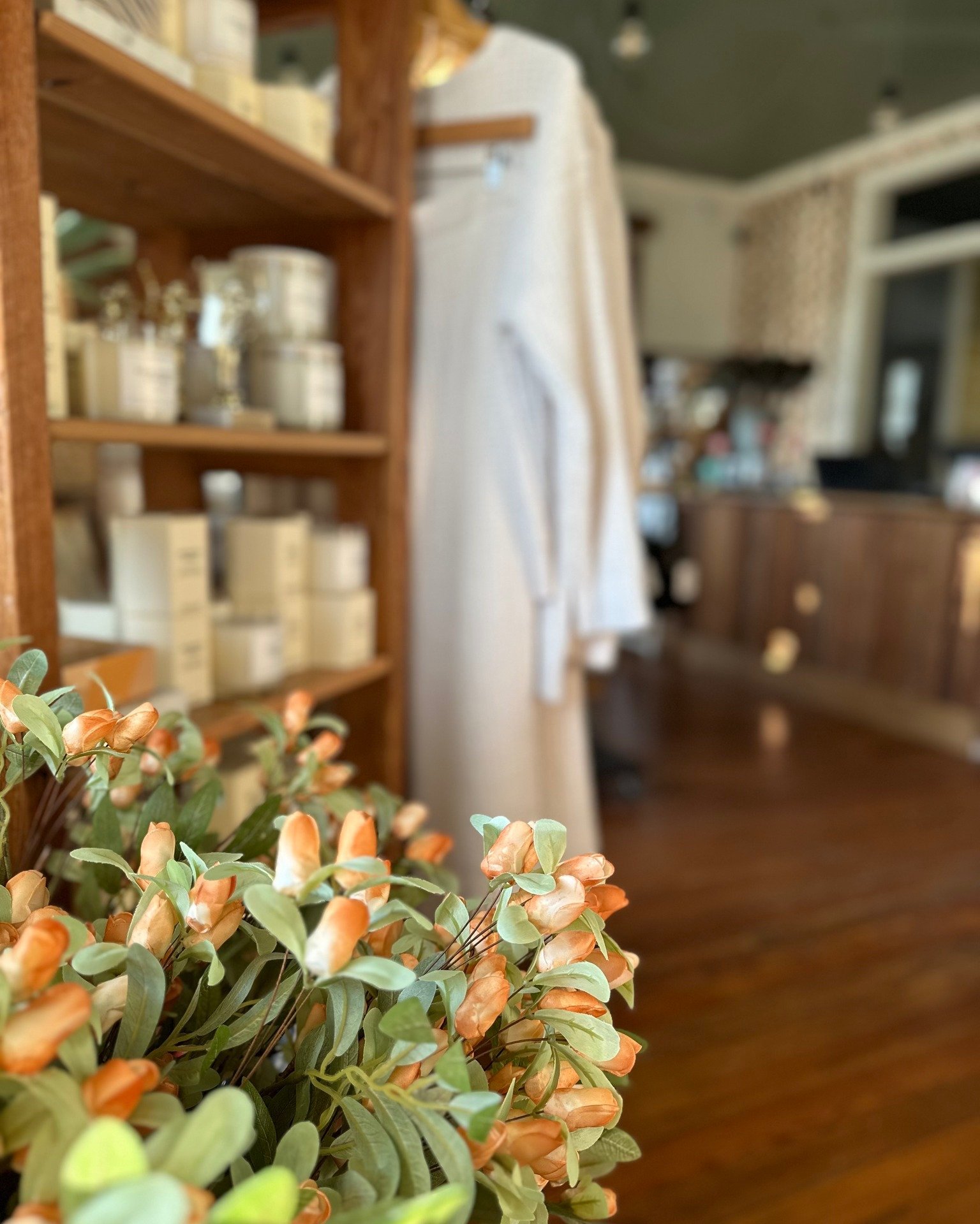 Close-up of orange and green flowers in the foreground, blurry interior of a store or cafe with wooden shelves and white robes hanging in the background.