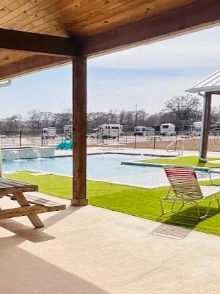 A covered patio area overlooking an outdoor swimming pool surrounded by a grassy area and a fence, with RV trailers and trees in the background.