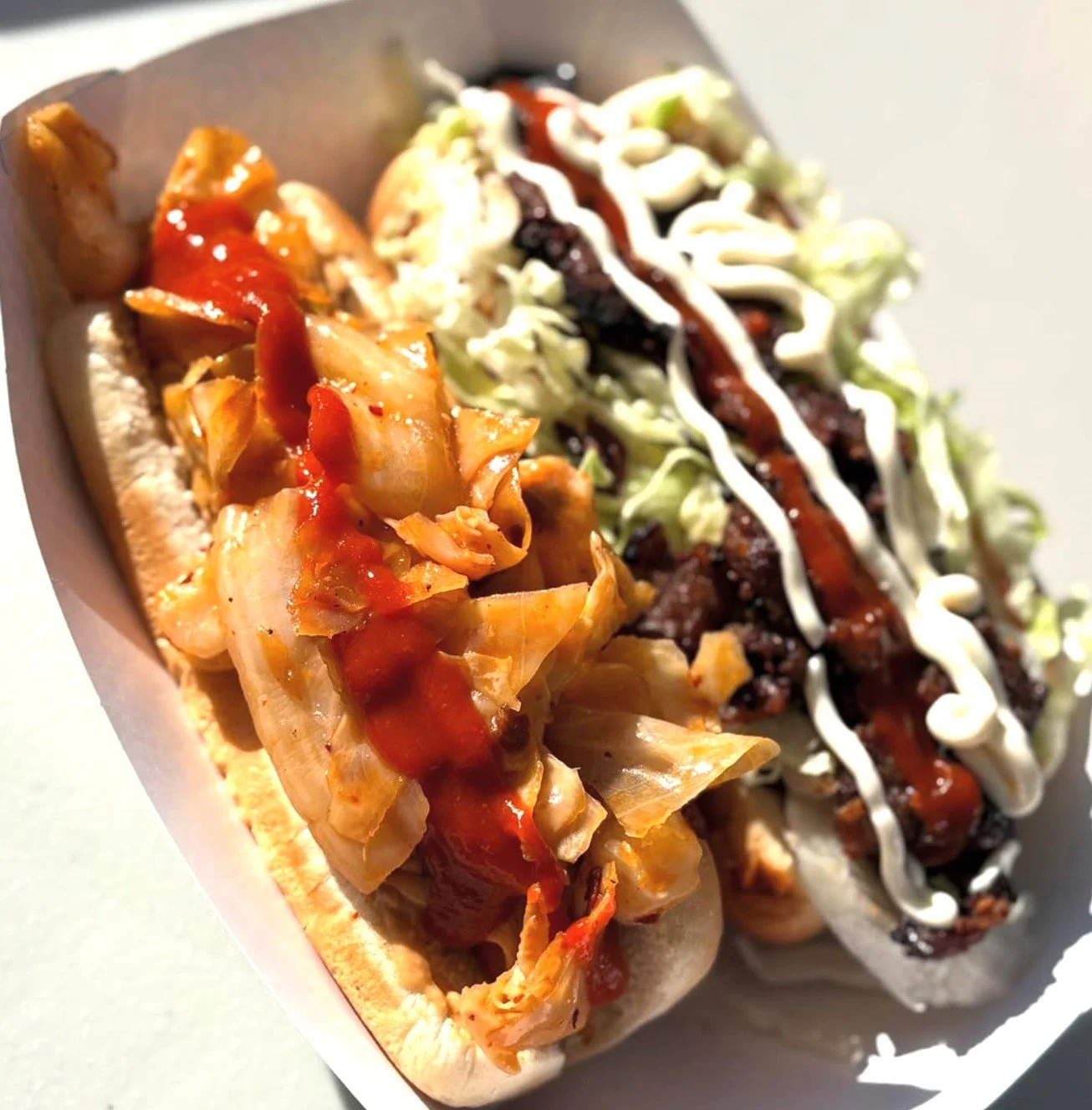 Two hot dogs with different toppings in a paper tray. The first hot dog has shredded chicken with tomato sauce, and the second has what appears to be seasoned ground beef with lettuce, mayonnaise, and barbecue sauce, topped with a zigzag of mayonnaise.