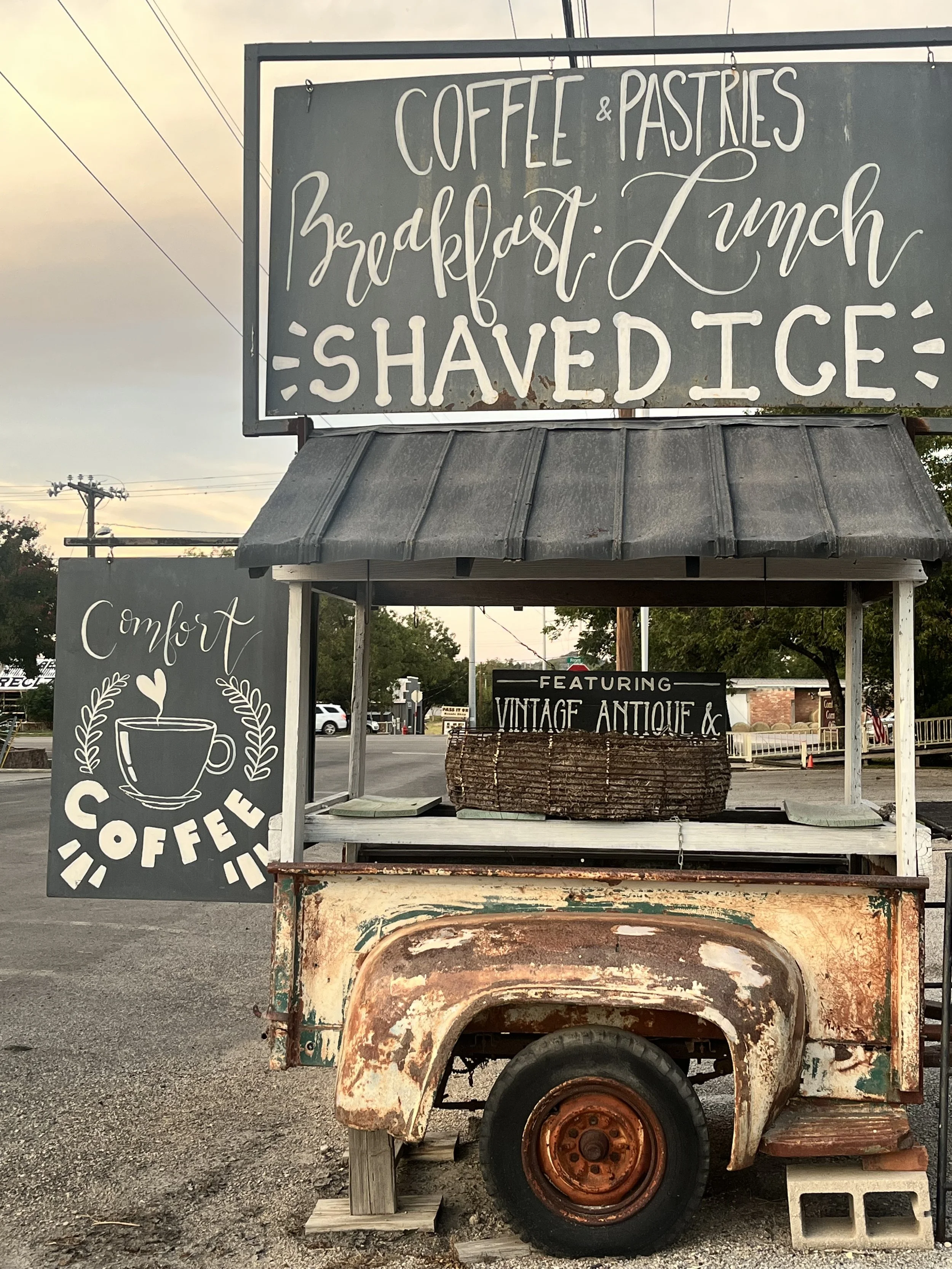 Vintage food cart with signs advertising coffee, pastries, breakfast, lunch, shaved ice, and vintage antique items, with a parking lot in the background.