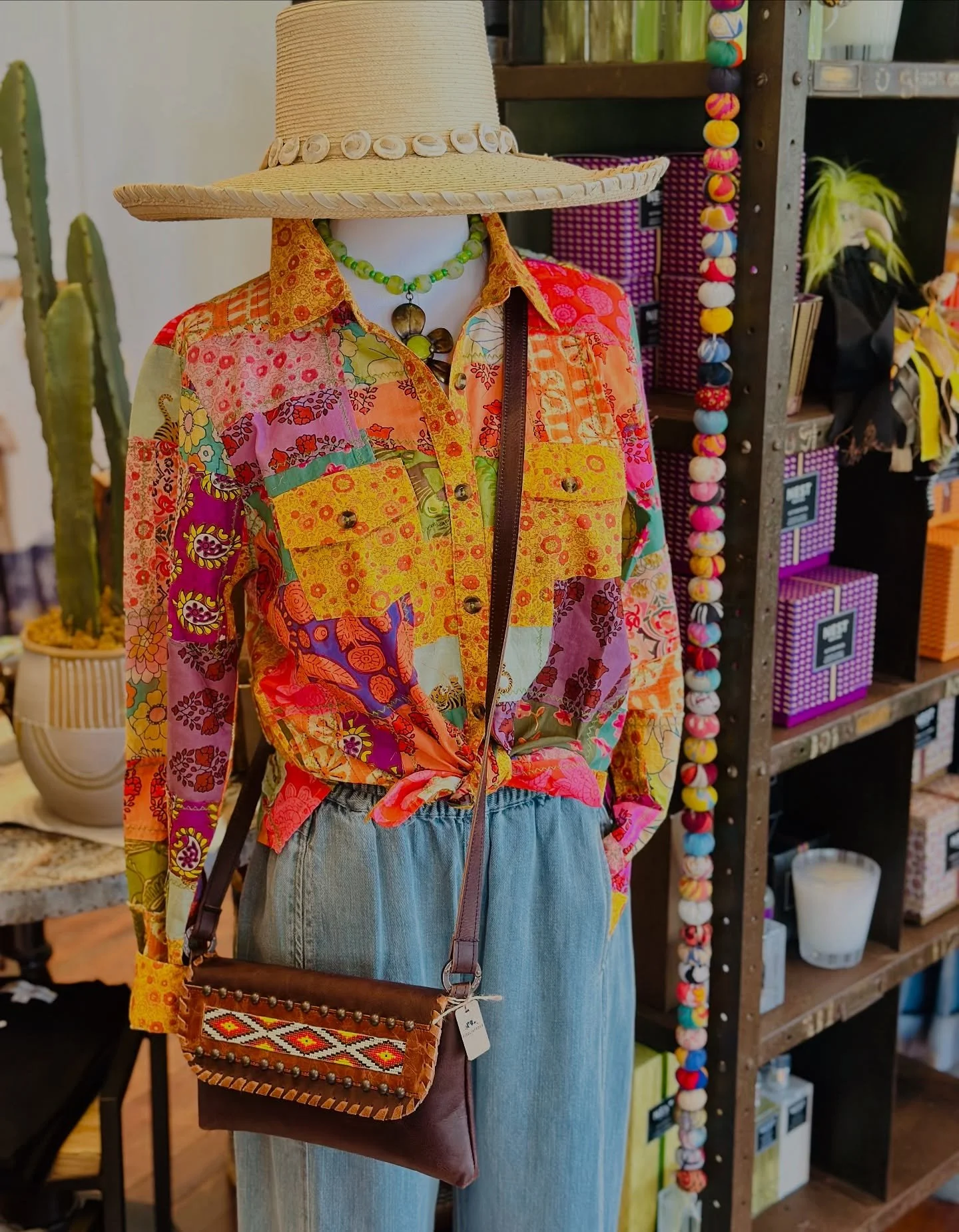 A mannequin wearing a brightly colored patchwork shirt, a wide-brimmed straw hat, a green beaded necklace, and light blue denim jeans, with a small embroidered crossbody bag.