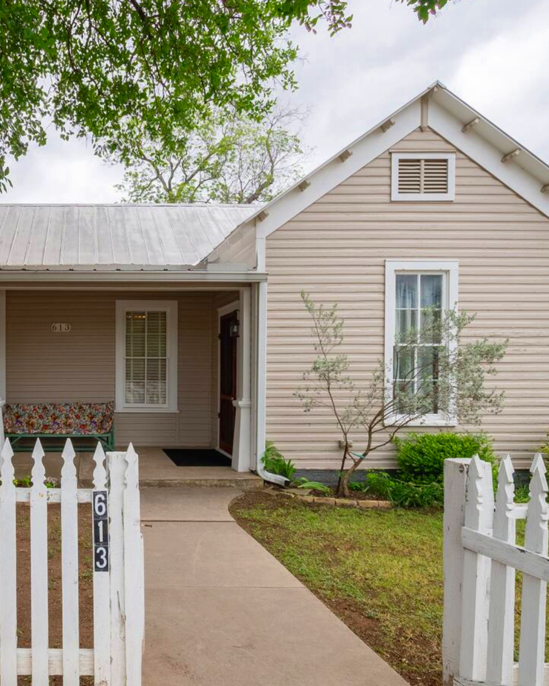 Front view of a beige house with white trim, a small front porch, and a white picket fence. The house has two windows and a gabled roof, surrounded by a small lawn and trees.