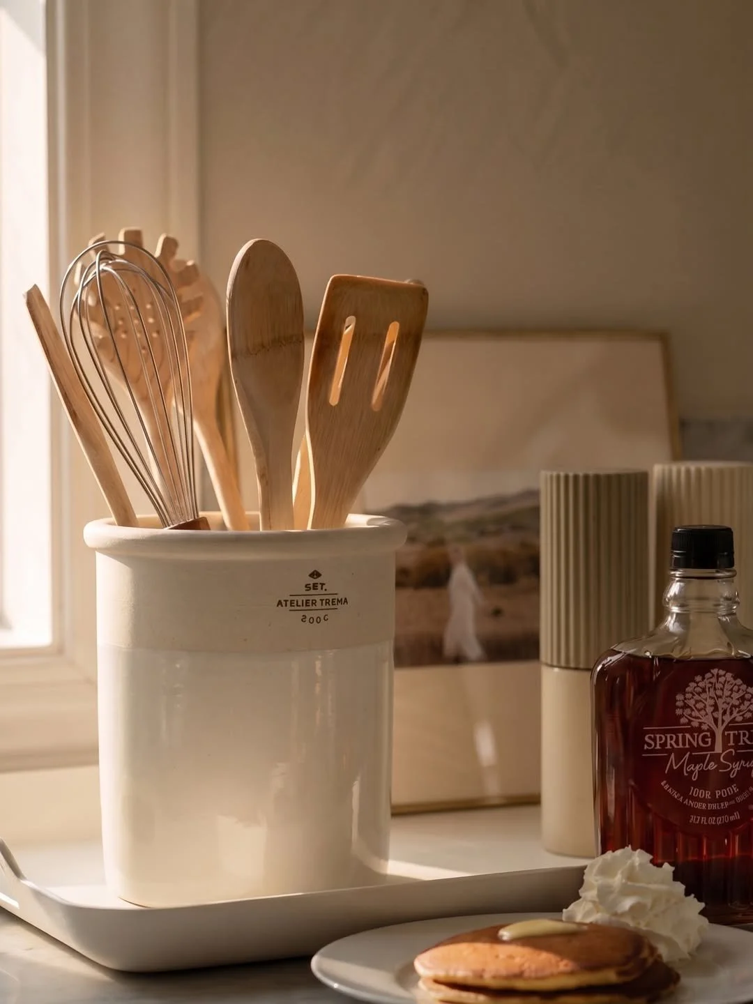 Wooden kitchen utensils in a white ceramic holder, a bottle of maple syrup, a plate with a pancake, whipped cream, and a small white dish, on a kitchen counter near a window with a landscape painting in the background.