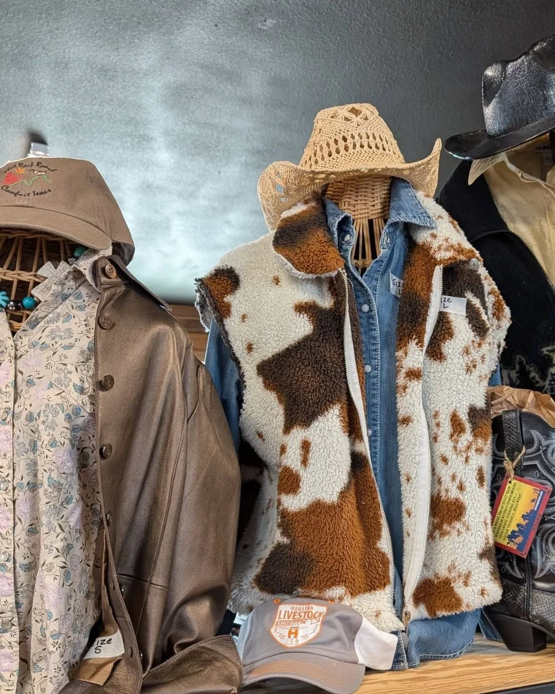 Display of cowboy and Western-style clothing on mannequins, including a cowhide patterned vest, denim shirt, cowboy hat, and leather jacket at a store.