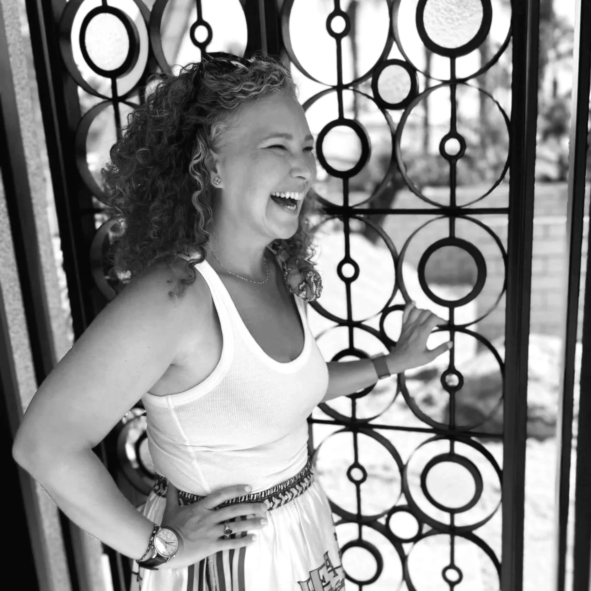 Kate smiling and laughing, standing in front of a decorative metal gate with circular patterns, outdoors in black and white.