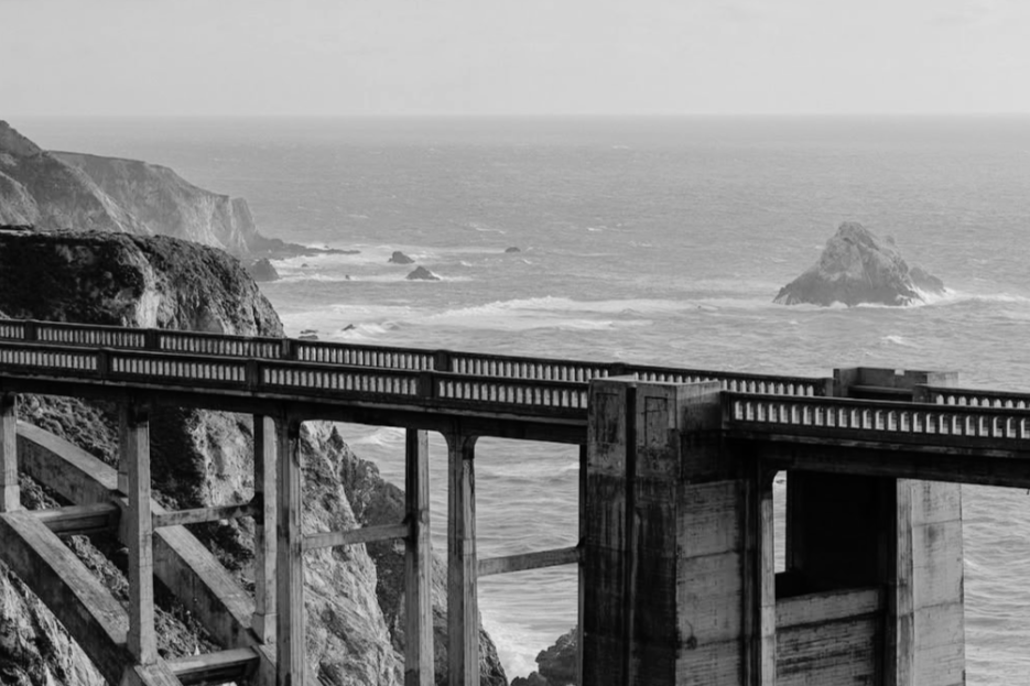 Black and white photo of the Bixby bridge over rocky cliffs and the ocean, with a large rock formation in the water in the background.