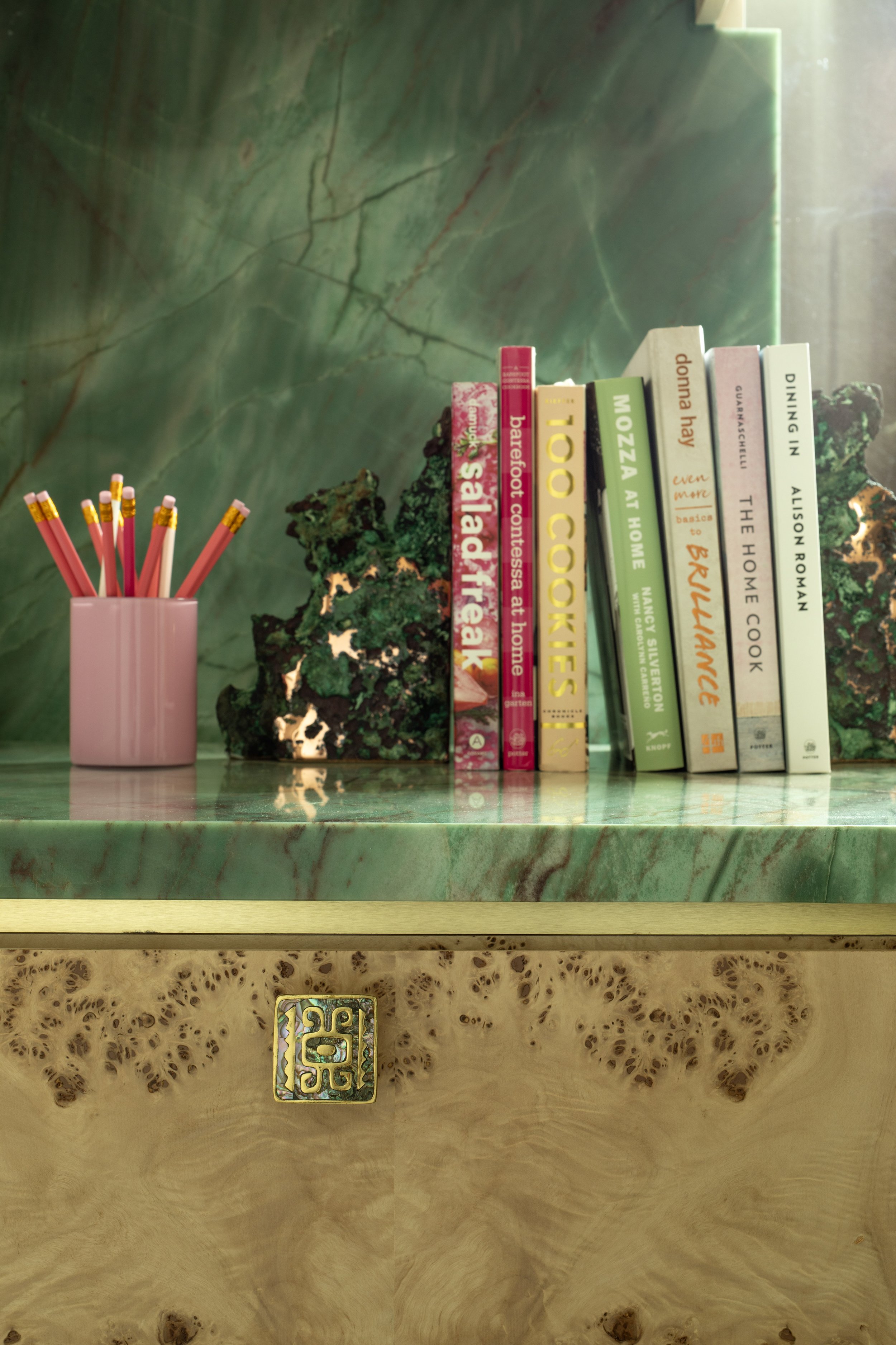 Pink pencil cup filled with red pencils on green marble surface next to decorative green rocks and a row of books about food, cooking, and home recipes.