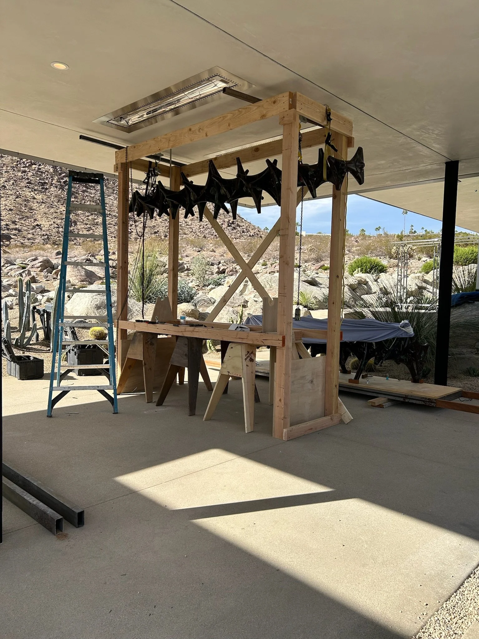 A wooden structure under construction holds multiple black megaphones hanging from chains, with a blue ladder and work tables around it. The setting appears to be an outdoor workshop space with desert landscape in the background.