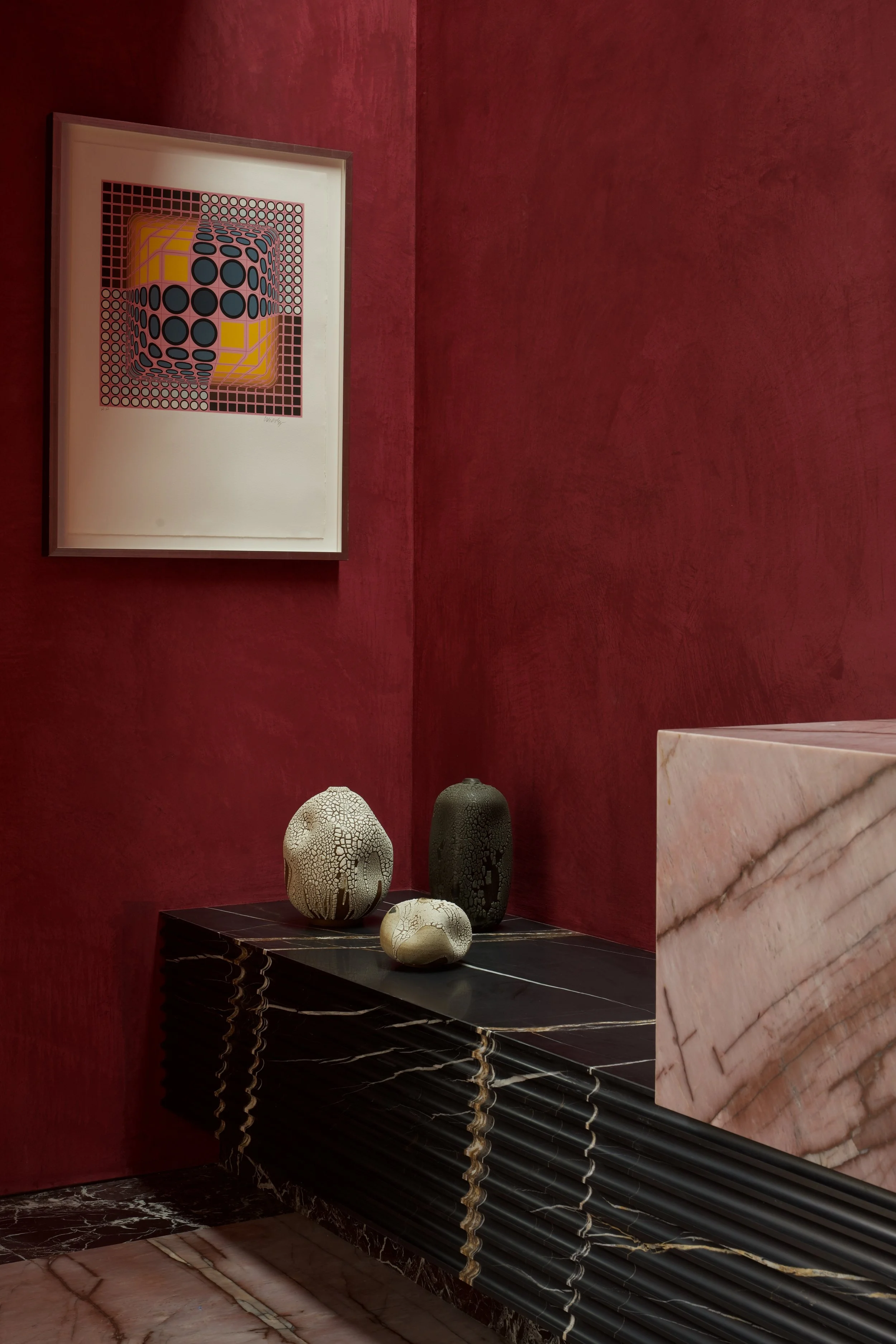 Decorative interior with red textured walls, a framed geometric artwork, three ceramic vases with intricate patterns on a black marble console, and a pink marble reception desk.