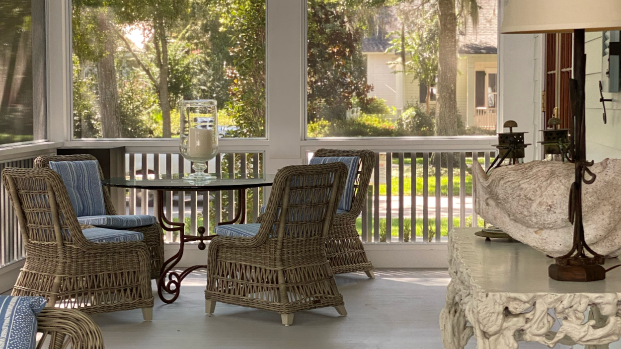 A sunlit screened porch with wicker chairs and a glass-topped table, overlooking a lush green yard with trees and houses in the background.
