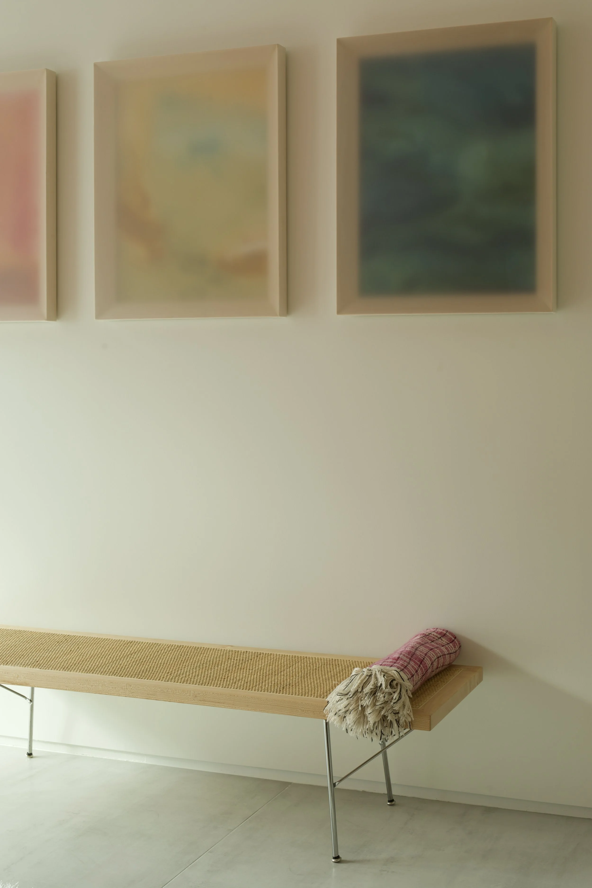 A minimalist interior with a wooden bench with thin metal legs and a pink checkered blanket rolled on one end, set against a light-colored wall decorated with three abstract, softly blurred framed artwork.