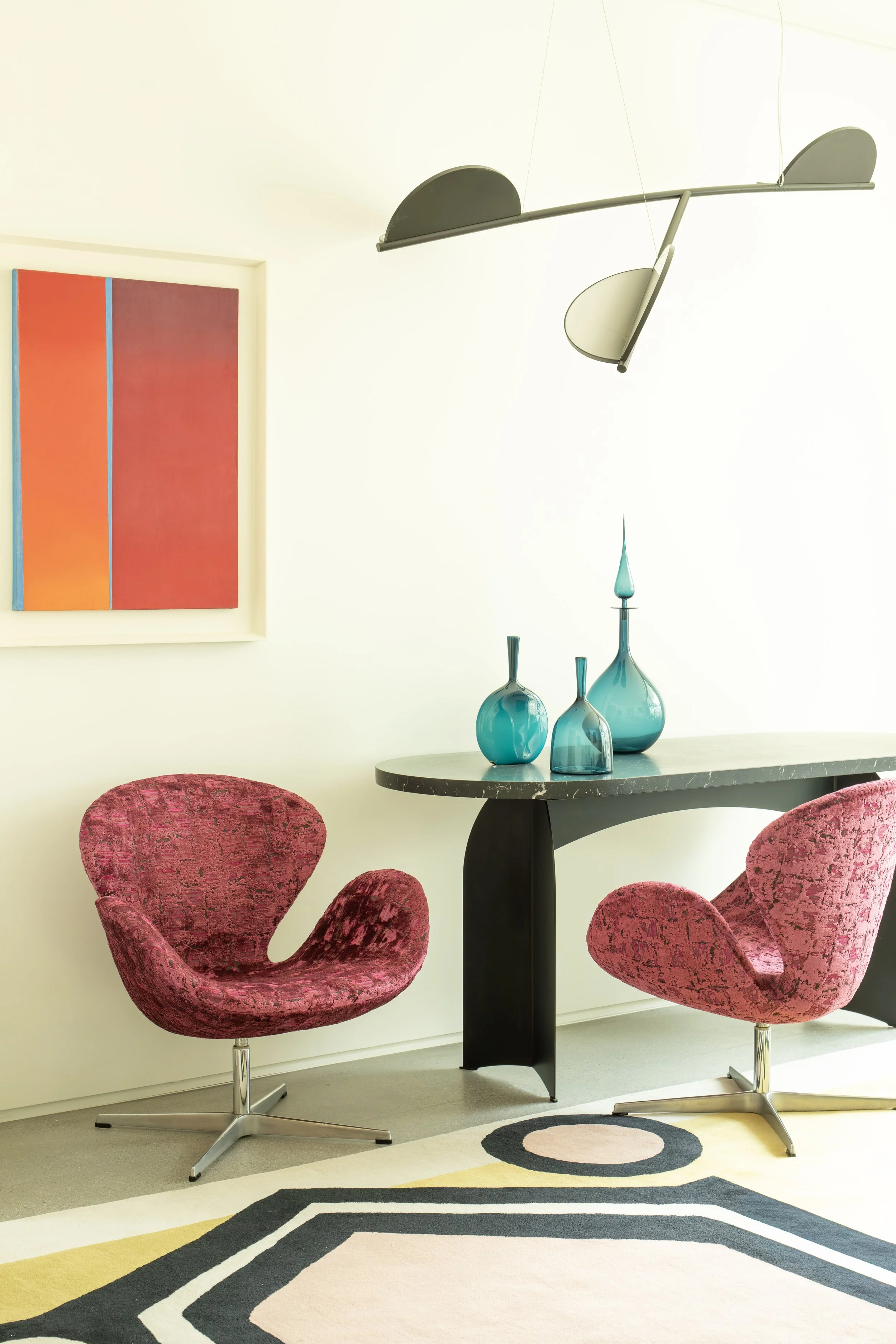 A modern dining room with two pink patterned chairs, a black marble table with three blue vases, a colorful abstract painting on the wall, a stylish black and white light fixture hanging from the ceiling, and a geometric black, white, pink, and yellow rug on the floor.