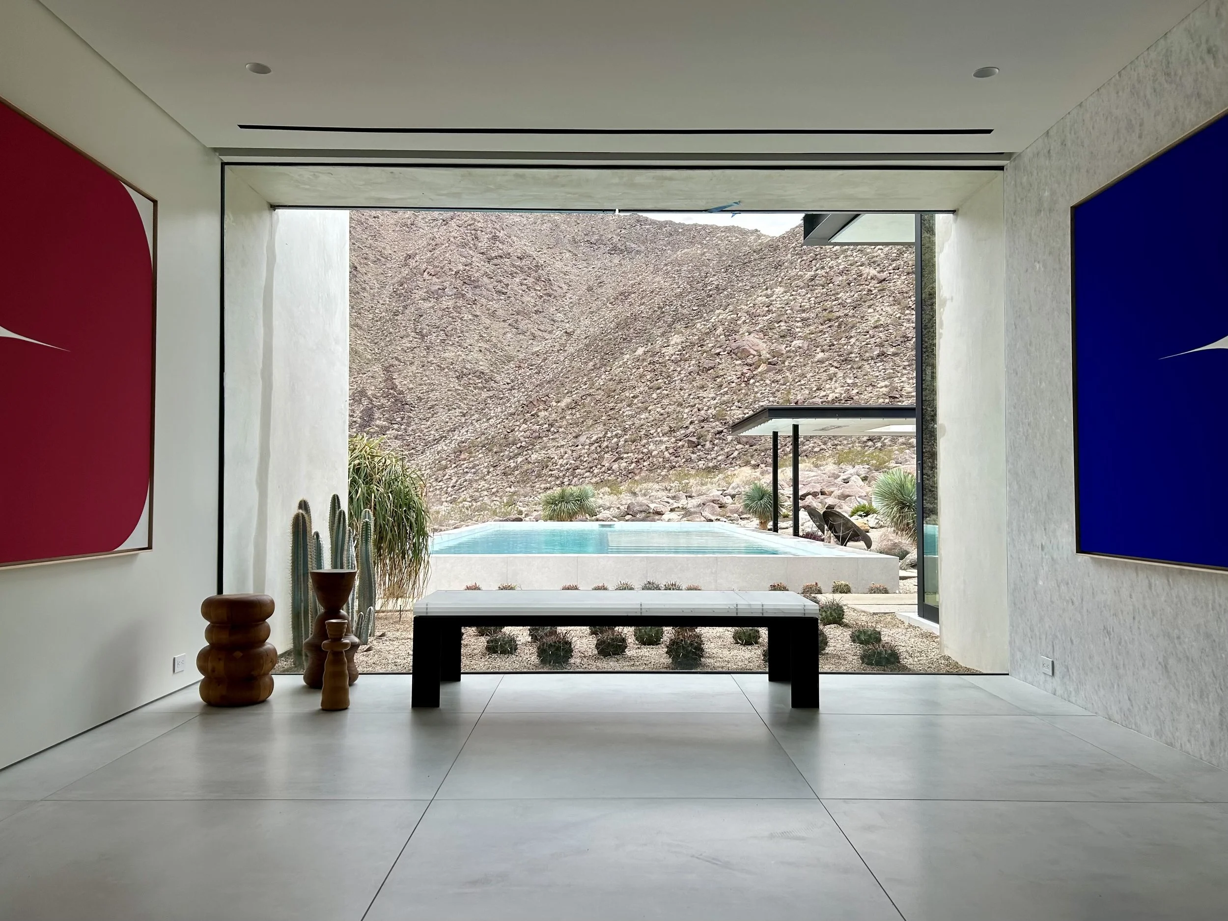 Modern interior room with minimal decor, featuring large window opening to desert landscape with mountains, pool, and cacti outside. Walls have colorful abstract art, and there is a black table in the center.
