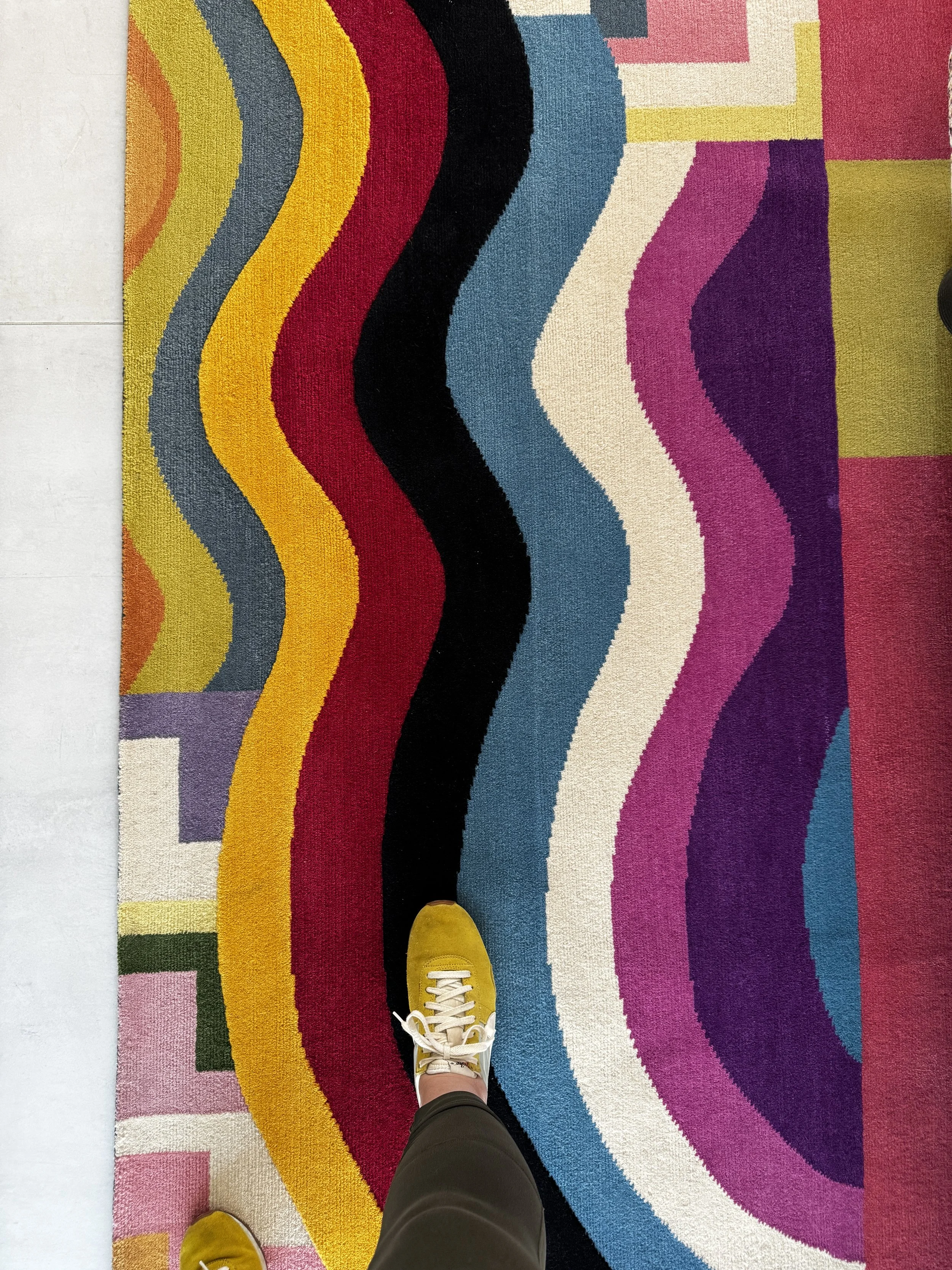 Top view of a colorful geometric patterned rug with a person's leg in black pants and yellow shoes standing on it.