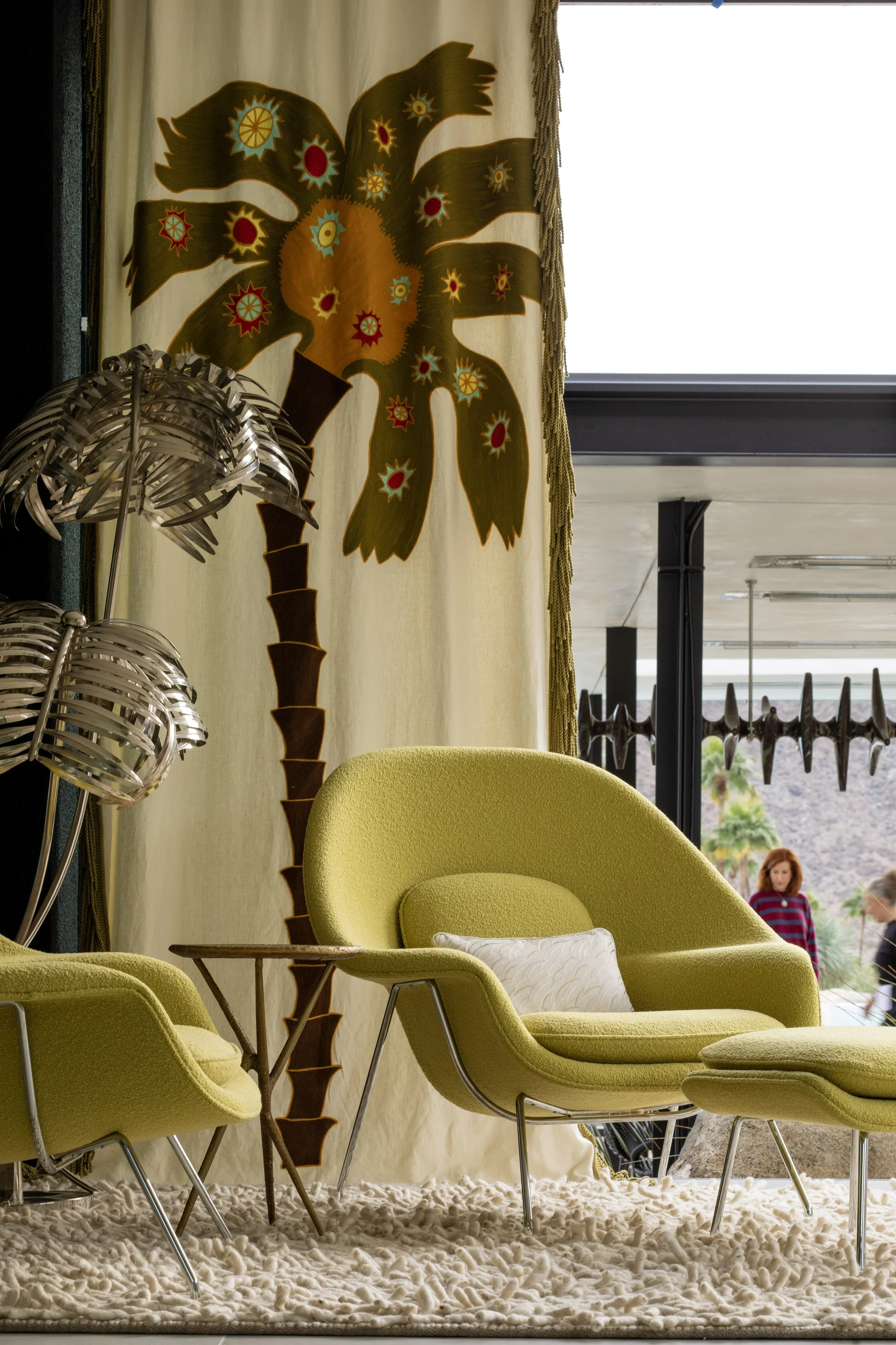 Interior with retro green lounge chairs, a silver palm leaf lamp, and a large fabric curtain with a stylized palm tree design.