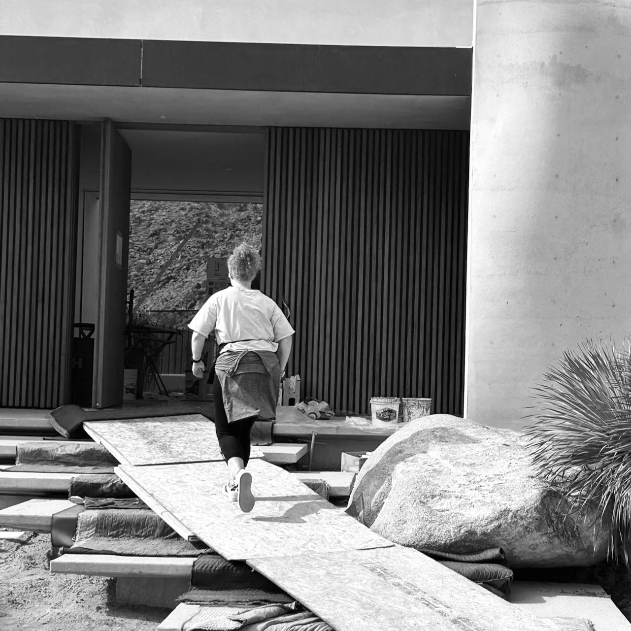 Kate, walking on a ramp towards an open building or studio with a modern exterior. There is a large rock and some plants nearby.